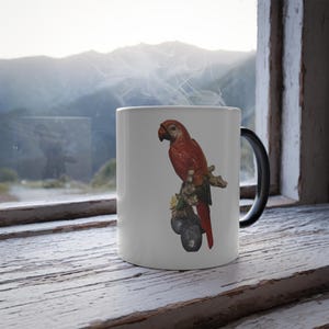 May include: White ceramic mug with a detailed illustration of a red parrot on a branch. The parrot has a red body and a black beak. The mug has a black handle and is set on a wooden surface. The background is white.
