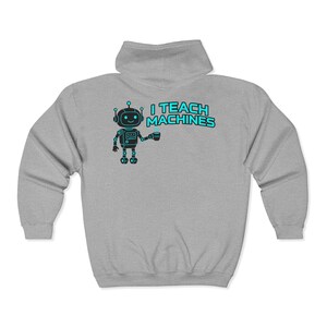 Könnte beinhalten: Hellgrauer Kapuzenpullover mit Reißverschluss und einem Cartoon-Roboter-Motiv mit dem türkisfarbenen Text "I TEACH MACHINES". Der Roboter hält eine Tasse. Der Hoodie hat eine Kapuze und einen durchgehenden Reißverschluss.