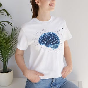 Könnte beinhalten: Weißes T-Shirt mit einem blauen Gehirn-Motiv, das ein Netzwerk aus leuchtenden Linien zeigt. Das Gehirn-Design ist von einem Spritzeffekt umgeben. Das Shirt hat kurze Ärmel und einen Rundhalsausschnitt.