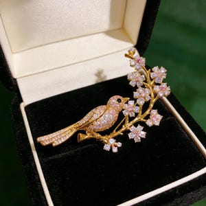 May include: A gold-coloured bird brooch with pink and clear crystal detailing, perched on a branch with crystal flowers. The bird's body is covered in pink crystals, and the flowers have pink centres. The brooch is presented in a black velvet box.