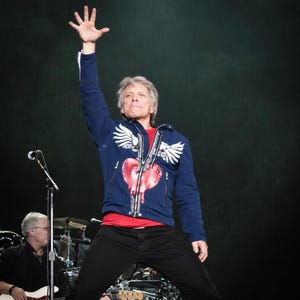 Op de afbeelding: Een man op het podium draagt een marineblauwe jas met witte vleugelontwerpen en een rode hartafbeelding. Hij draagt een rood shirt en een zwarte broek, met zijn arm omhoog. Een microfoon en bandleden zijn zichtbaar op de achtergrond.
