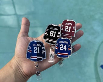 May include: Four hockey jersey-shaped badge reels with glitter borders. The jerseys feature names and numbers: Rozanov 81, Hunter 21, Hollander 24, and a blue jersey with 21. Each has a retractable cord and a clear plastic strap attachment.