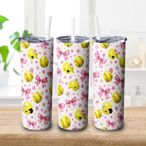 Peut inclure: Trois gobelets isothermes blancs avec couvercles et pailles. Chaque gobelet présente un motif répété de balles de softball jaunes, de casques, de nœuds roses et de fleurs blanches sur fond blanc. Les gobelets sont sur une surface en bois.
