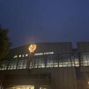 以下が含まれることがあります： 夕暮れ時の帯広駅の外観。大きな時計と白い文字で「OBIHIRO STATION」と書かれた建物。空は深い青色です。