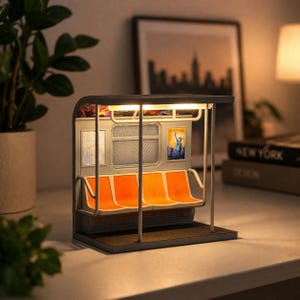 May include: A miniature New York City subway car lamp with orange seats, a light-up interior, and a poster of the Statue of Liberty. The lamp has a black roof and a gray exterior, with a base that sits on a table.