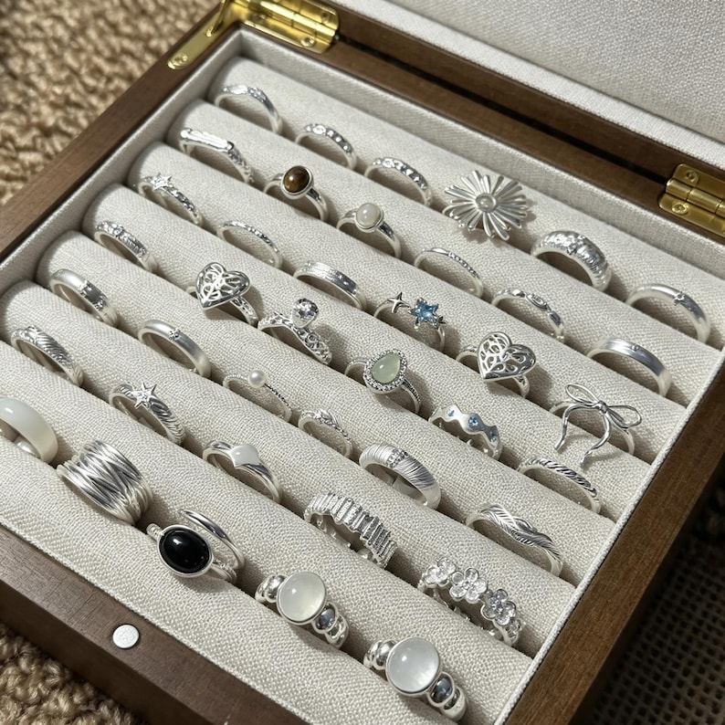 May include: A collection of silver-toned rings arranged in a wooden jewelry box with a light-colored velvet interior. The rings feature various designs including heart shapes, floral motifs, smooth bands, textured surfaces, and inset stones.