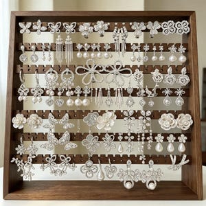 May include: A dark wood jewelry display stand featuring multiple horizontal rows with notched edges, holding a large collection of silver-tone earrings. The earrings include various designs such as floral shapes, dangling pearls, crystals, and butterflies.