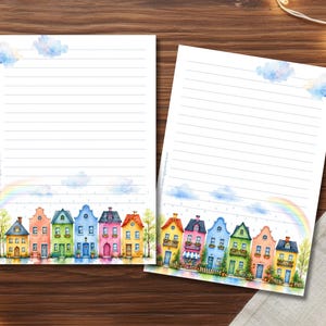 Könnte beinhalten: Zwei Blätter liniertes Briefpapier mit einer Aquarellillustration von bunten Häusern, einem Regenbogen und Wolken. Die Häuser sind in verschiedenen Farben gehalten, darunter Blau, Rosa, Gelb und Grün. Das Briefpapier liegt auf einer Holzoberfläche.