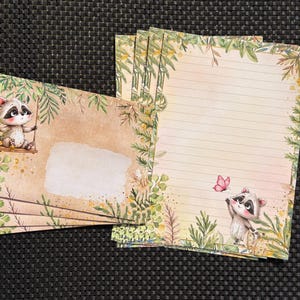Könnte beinhalten: Ein Schreibwarenset mit einem Waschbären auf einer Schaukel auf den Umschlägen und einem Waschbären, der nach einem rosa Schmetterling greift, auf dem linierten Papier. Beide Artikel haben einen beigen Hintergrund mit grünen Blattbordüren.