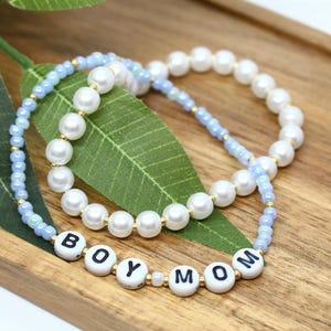 May include: Two beaded bracelets. One bracelet features white pearl beads and gold accents. The other bracelet has light blue beads and the letters "BOY MOM" spelled out on white beads. The bracelets rest on a wooden tray.