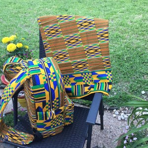 May include: A colorful backpack and a matching blanket rest on a black chair outdoors. The backpack and blanket feature a vibrant geometric pattern in yellow, blue, green, and orange. The scene is set against a backdrop of green grass and foliage.