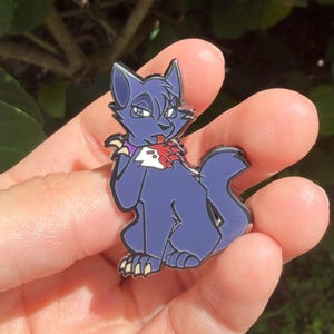 Puede incluir: Un pin de esmalte azul oscuro que muestra un gato estilizado con orejas puntiagudas, un collar morado y una mancha blanca en el pecho. El gato aparece sentado con una pata levantada hacia la boca, mostrando garras afiladas y un detalle rojo.