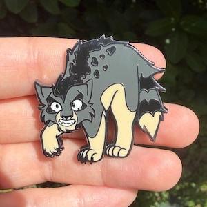 Op de afbeelding: Een metalen emaille speldje met een gestileerd cartoonwezen met grijze en cr&egrave;mekleurige vacht, een zwart gestreepte rug en een grimmige uitdrukking. Het figuurtje heeft puntige oren, scherpe tanden en een gebogen houding met een pluizige staart.