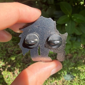 Op de afbeelding: De achterkant van een metalen emaille speld met een getextureerde, donkergrijze glitterafwerking. Twee ronde zwarte rubberen sluitingen zijn aan de achterzijde bevestigd en beveiligen de twee metalen pinnen voor bevestiging aan kleding.