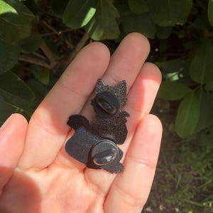 Puede incluir: Un pin de esmalte negro con un acabado texturizado y brillante en el reverso. El pin tiene dos cierres circulares de goma negra sujetos a los postes met&aacute;licos, los cuales est&aacute;n posicionados en la parte superior e inferior de la estructura.