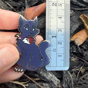 Puede incluir: Un pin de esmalte azul oscuro con forma de gato de dibujos animados con garras amarillas y un collar morado, junto a una regla de metal que muestra una altura de unos 4,5 cent&iacute;metros. El gato tiene una marca roja en el pecho.