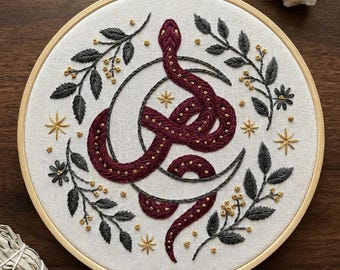 May include: Embroidered hoop art featuring a burgundy snake coiled around a crescent moon. The design includes gold-colored stars and accents, with dark gray leaves and gold-colored berries. The embroidery is set in a wooden hoop.