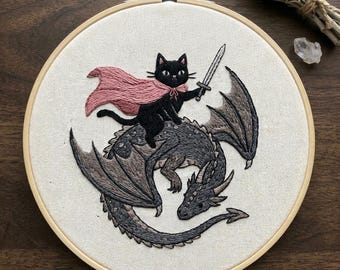 May include: Embroidered hoop art showing a black cat in a pink cape, wielding a sword, and riding a grey dragon. The design is on cream fabric, framed by a wooden embroidery hoop. The dragon has detailed wings and scales, a fantasy theme.