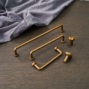 May include: A collection of antique brass cabinet hardware, including bar pulls and round knobs. The hardware is arranged on a dark wood surface, with a soft, gray fabric in the background. The pulls vary in size and shape.