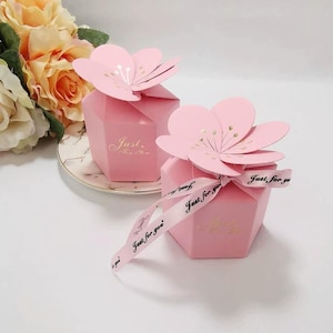 Puede incluir: Dos cajas de regalo hexagonales rosas con tapas en forma de flor. Las cajas tienen texto dorado que dice "Just for you". Una caja está atada con una cinta que también dice "Just for you."