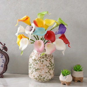 Puede incluir: Un arreglo floral con calas coloridas en un jarrón decorativo. Las flores muestran una variedad de tonos, incluyendo amarillo, rojo y morado. El jarrón tiene un patrón floral. También hay dos pequeñas plantas suculentas en macetas decorativas.