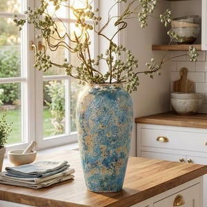 May include: A tall, textured blue and tan ceramic vase filled with green-berried branches sits on a wooden countertop. The vase is the focal point, with a rustic, aged appearance. A window and other kitchen items are in the background.