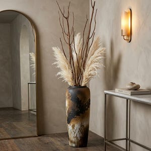 May include: A tall, decorative vase with a black, brown, and white mottled finish. The vase is filled with dried pampas grass and brown branches. The vase is placed near a mirror and a side table.