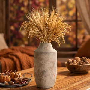 May include: A tall, textured, gray vase holds a large bundle of dried wheat stalks. The vase sits on a wooden table, with a tray of pumpkins, acorns, and cinnamon sticks in the foreground. A bowl of nuts is also visible.