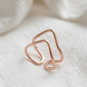 May include: A rose gold ring featuring a double-layered, open-ended design with smooth, rounded edges. The band consists of two parallel, wave-like shapes that create a geometric, minimalist silhouette, showcasing a polished metallic finish.