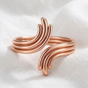 May include: A rose gold-toned open ring featuring a multi-strand design. The band consists of three parallel wires that curve upward at both ends, creating a layered, fan-like appearance. The metal has a smooth, polished, and reflective finish.