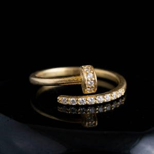 Puede incluir: Un anillo dorado con un diseño de cabeza de clavo. El anillo presenta una fila de pequeñas piedras preciosas transparentes a lo largo de la banda y en la cabeza del clavo. El anillo está sobre un fondo negro, creando un contraste llamativo.