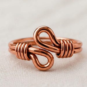 Peut inclure: Une bague en cuivre fabriquée à partir d'un fil continu, présentant deux boucles symétriques au centre et deux sections de fil étroitement enroulées autour de l'anneau de chaque côté des éléments de conception centraux.