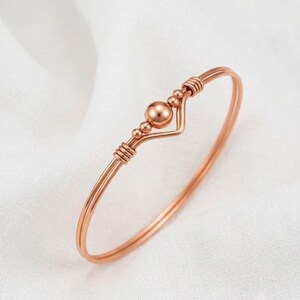 May include: A rose gold-toned wire bangle bracelet featuring a central spherical bead flanked by two smaller beads. The wire forms a V-shaped chevron detail at the centre, with coiled wire accents securing the beads to the main circular band.