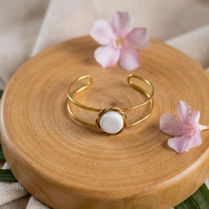 Könnte beinhalten: Ein goldfarbenes Metallarmband mit einem weißen, kreisförmigen Mittelstück. Das Armband hat ein Doppelband-Design und liegt auf einer Holzoberfläche mit rosa Blüten.