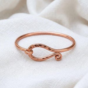 Peut inclure: Un bracelet jonc en cuivre présentant une texture martelée et un mécanisme de fermeture central de type crochet. Le métal possède une finition métallique brun-rouge chaleureuse avec une surface polie qui reflète la lumière sur la forme.