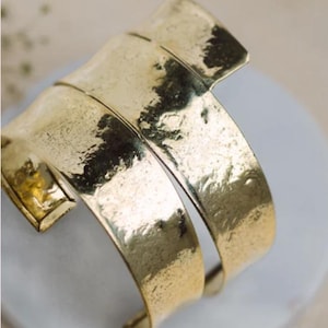 May include: A gold-colored cuff bracelet with a textured surface. The bracelet has a wide, open design with a curved shape. The metal has a hammered appearance, giving it a rustic, yet elegant look.