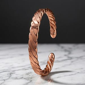 Pode incluir: Uma pulseira tipo bracelete de cor cobre com um design contínuo de corda torcida. O metal tem um acabamento fosco escovado com reflexos sutis ao longo das cristas do padrão em espiral. A pulseira é aberta com extremidades arredondadas.