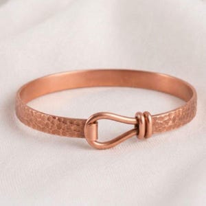 May include: A copper-colored metal cuff bracelet featuring a hammered, textured surface throughout the band. The bracelet is designed with a central hook-and-loop style closure mechanism that secures the two ends of the circular metal band together.
