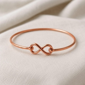 Può includere: Un bracciale rigido in metallo color oro rosa con un simbolo dell'infinito al centro. Il sottile cerchio è lucido e liscio, con l'anello dell'infinito integrato direttamente al centro della struttura del bracciale.