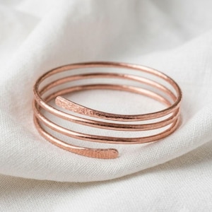 May include: A copper-colored metal bangle bracelet featuring a multi-coil spiral design. The wire has a textured, hammered finish along its surface, with two tapered, flattened ends that complete the circular, layered wrap-around structure.