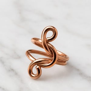 May include: A polished copper-toned ring featuring a continuous, fluid wire design that loops and curves into an open, abstract shape. The metal surface is smooth and reflective, showcasing a warm, metallic hue throughout the entire structure.