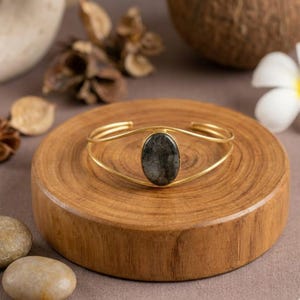 Può includere: Un bracciale a polsino in metallo color oro con una pietra ovale grigio scuro al centro. Il bracciale è esposto su una superficie rotonda di legno. Lo sfondo include elementi naturali come pietre e fiori secchi.