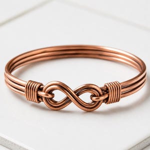 Può includere: Un bracciale rigido in filo di rame caratterizzato da un design a tre fili che convergono in un simbolo dell'infinito centrale. Le due estremità del bracciale sono fissate con avvolgimenti stretti di filo di rame, creando un anello continuo.