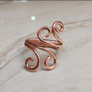 May include: A copper-colored wire ring featuring a central band wrapped with thin wire coils and two symmetrical, upward-curving scroll designs on each side. The metal has a polished, metallic finish with a warm, reddish-brown hue throughout.