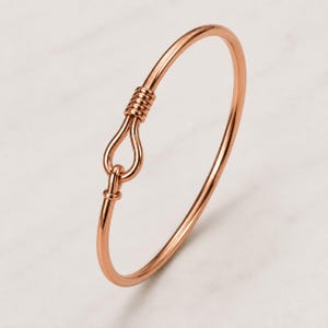 Pode incluir: Uma pulseira rígida em tom de ouro rosa com uma banda circular lisa que termina num fecho de gancho. O mecanismo de fecho consiste numa extremidade em forma de laço, fixada por uma série de espirais de arame apertadas na superfície.