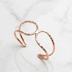 May include: A rose gold cuff bracelet featuring two interconnected open oval shapes. The metal surface has a hammered texture, creating a series of small indentations along the thin, curved bands that form the structure of this open-ended wrist accessory.