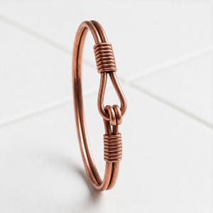 May include: A copper wire bracelet featuring a circular band design with a central hook-and-eye closure mechanism. The metal is formed into two parallel strands that are secured together by tightly coiled copper wire wraps at the clasp area.