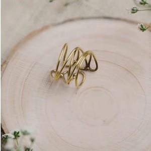 Könnte beinhalten: Ein goldfarbener, abstrakter Ring mit einem Spiraldesign. Der Ring besteht aus dünnem, gebogenem Metall und liegt auf einer hellen Holzoberfläche. Das Design des Rings weist mehrere Schlaufen und Kurven auf und erzeugt eine einzigartige und moderne Ästhetik.