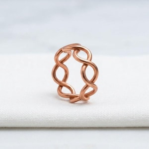 May include: A copper-coloured ring featuring a continuous, repeating twisted wire design. The band is constructed from a single strand of metal formed into a series of interconnected loops, creating an open, airy structure with a smooth, polished finish.