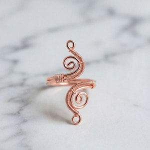May include: A copper wire ring featuring an open design with two symmetrical, coiled spiral ends. The metal wire is tightly wrapped around the base of each spiral, creating a textured, metallic finish that forms a continuous, adjustable band.